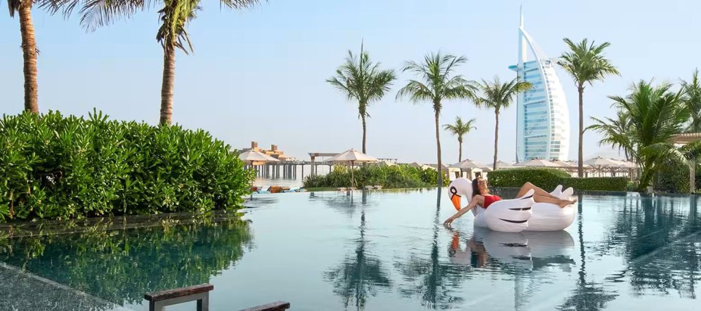 Dubaj. Kąpiel w basenie pod palmami. W tle słynny hotel o kształcie żagla.